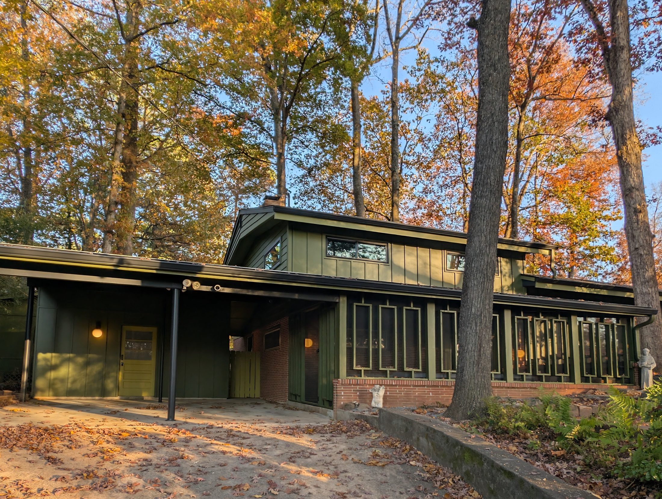 The front of the house in autumn: dark green board and batten siding over a brick base, a carport on slim steel posts, and a long run of geometric window frames under tall hardwoods in orange and gold
