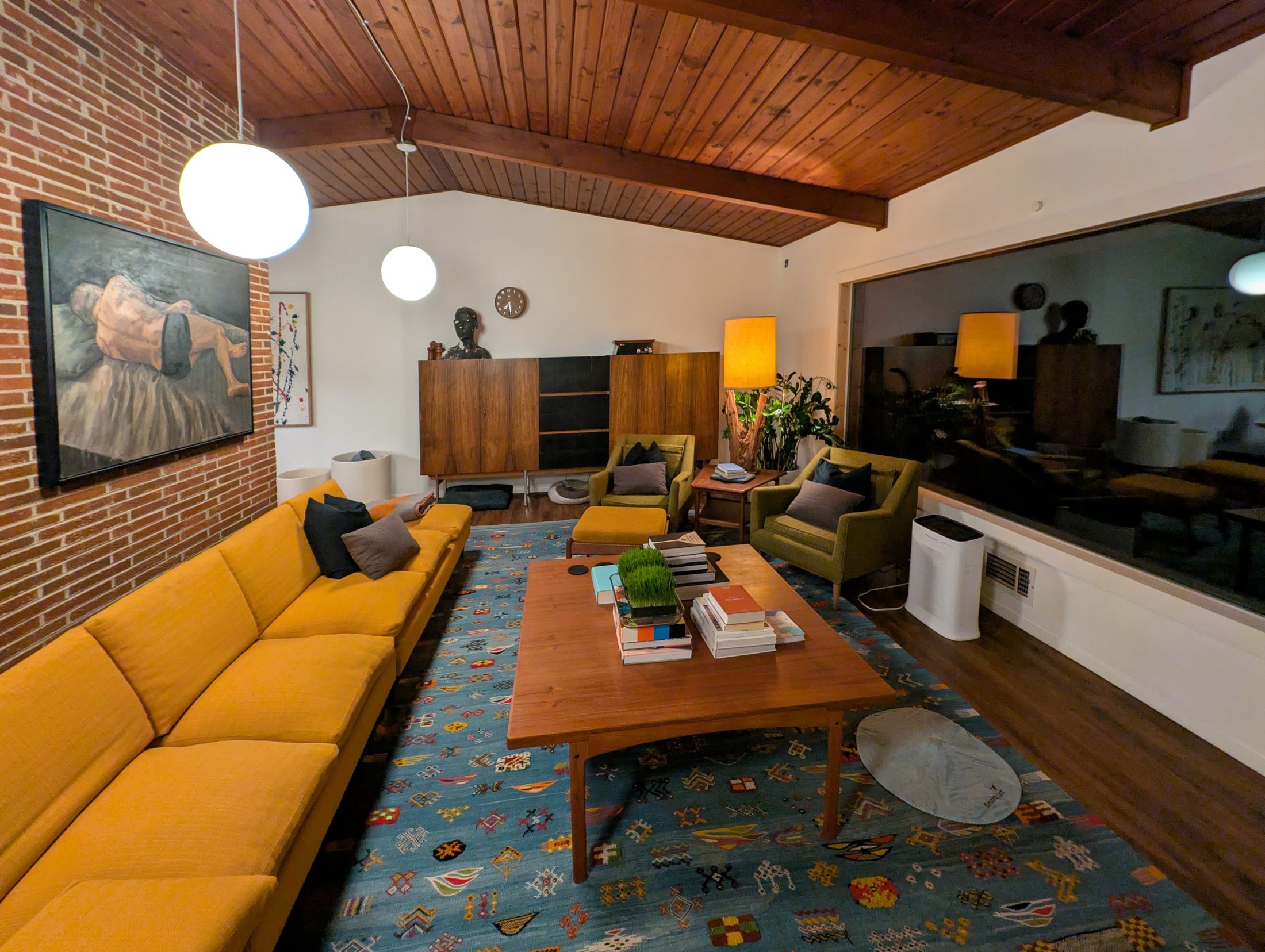 The living room: a warm wood plank ceiling on dark beams, a full brick wall, globe pendant lights, a long mustard sofa, walnut credenza and a patterned blue rug